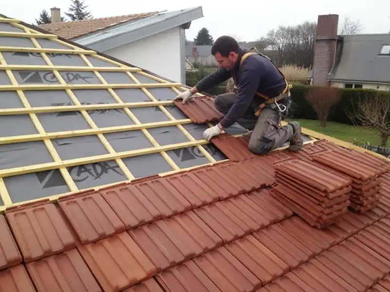 Un homme portant des &eacute;quipements de s&eacute;curit&eacute; est agenouill&eacute; sur un toit en pente, posant des tuiles d'argile rouge lors d'une r&eacute;novation de toiture dans le 91. Des piles de tuiles se trouvent &agrave; c&ocirc;t&eacute; de lui, des lattes de bois sont visibles, des maisons et des arbres apparaissent &agrave; l'arri&egrave;re-plan.